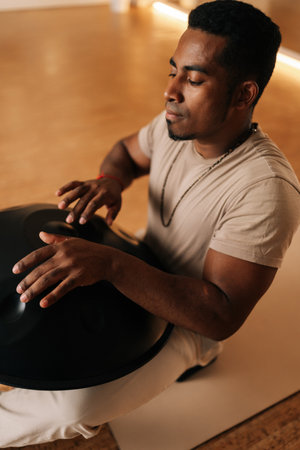 High-angle portrait of relaxed African man with closed eyes playing traditional ethnic music using glucophone sitting on floor in lotus pose. Serene black male drumming rhythm on tank drum by hands.の写真素材