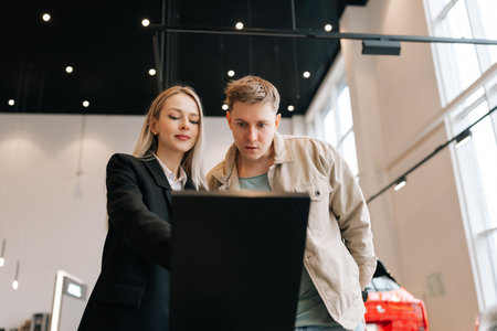 Low-angle view of successful woman seller consulting in auto showroom help male customer choosing new modern electric automobile. Professional saleswoman consult explain vehicle features sale.の写真素材