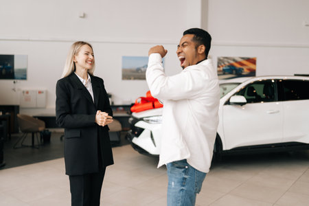 Happy excited African-American customer celebrating successful car purchase, dancing and having fun at auto dealership, enjoying new vehicle at automobile showroom. Concept of buying new auto.の写真素材