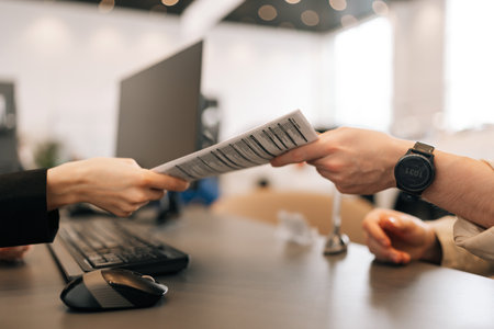 Close-up hands of unrecognizable manager and client exchanging document sitting at desk in office....