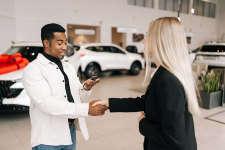 Happy satisfied African car owner getting key from smiling female auto dealer at dealership. Cheerful black male taking key to bought car and sealing deal with handshake in auto showroom.の写真素材