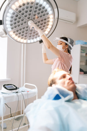 Vertical selective focus shot of female nurse in medical uniform adjusting light preparation performing medical procedure at clinic. Surgeon adjusting surgical lamp before medical operation.の写真素材