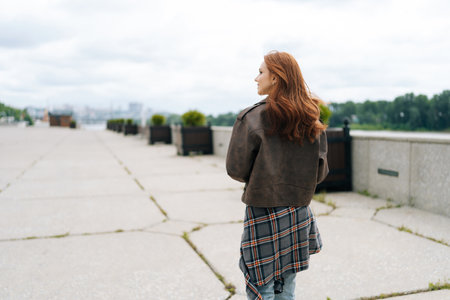 Back view of happy young woman with flowing red hair strolling along riverbank. Cheerful redhead female dressed in brown leather jacket, enjoying fresh air and beautiful scenery surrounding her.の写真素材