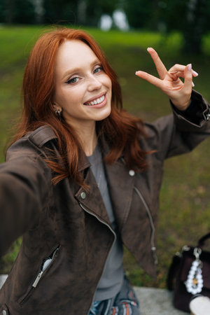 Vertical portrait of positive young woman with long red hair smiling happily looking at camera, taking selfie in park, showing peace sign. Happy redhead female shooting selfie flashing peace sign.の写真素材