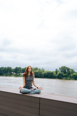 Full length vertical portrait of serene woman meditating with closed eyes sitting on wooden pier by river, enjoying fresh air and tranquility. Calm female practicing mindfulness, finding inner peaceの写真素材