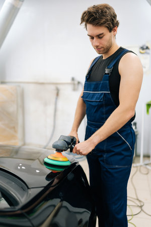 Vertical portrait of muscular auto mechanic worker using grinding unit to polish black hood at automobile repair and renew service station. Professional repairman in uniform polishing hood of car.の写真素材
