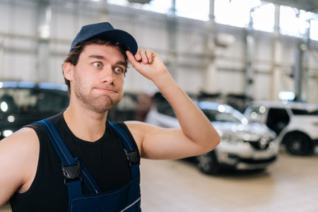 Closeup face of pensive thoughtful male mechanic in uniform holding visor of his cap with his hand puzzled looking away in auto service shop. Concept of professional automobile maintenance.の写真素材