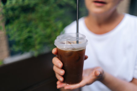Cropped shot of unrecognizable woman in white t-shirt holding takeaway plastic cup with cold coffee drink outdoors. Cup of iced bubble tea with straw in female hand. Concept of drink, city life.の写真素材