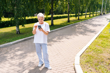 Full length portrait of senior aged woman typing on smartphone while walking in city park. Beautiful...