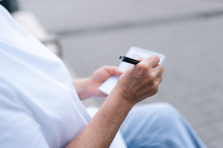 Closeup cropped shot of unrecognizable senior woman taking notes sitting on city bench, jotting down...