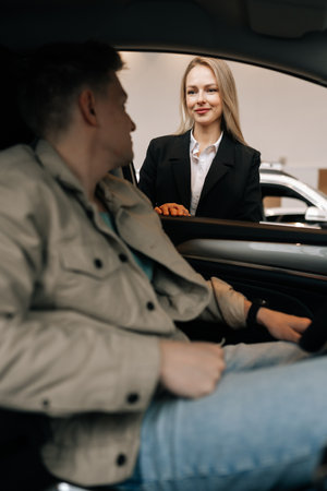 Vertical view from vehicle interior to confident female manager in suit talking to male client sitting in car in showroom choosing automobile. Concept of choosing and buying new auto at showroom.の写真素材