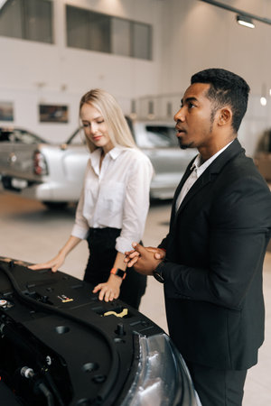 Vertical shot of African salesman standing near luxury car with open hood providing details review for blonde female customer at salon. Businesswoman choosing vehicle at showroom with help of manager.の写真素材