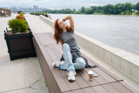 High-angle view of serene pretty young woman sitting on wooden platform by river, enjoying peaceful moment with coffee, cloudy sky and calm water creating serene atmosphere.の写真素材