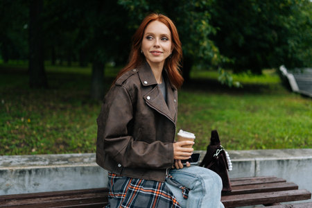 Portrait of fashionable young female in leather jacket enjoying free time with savors tranquil...