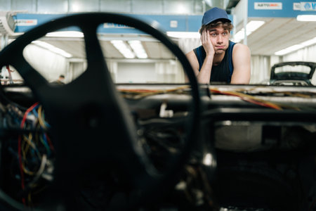 Tired mechanic fixing car in repair garage, frustrated but focused, surrounded by tools and equipment. Pensive repairman in car service looking at car thinking of how to repair in garage car service.の写真素材