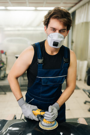 Vertical portrait of mechanic in face mask polish hood using grinding unit in automobile repair and renew service station. Repair after an accident. Concept of car repair and maintenance work.の写真素材