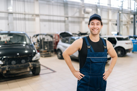 Portrait of cheerful male repairman in uniform standing posing in auto service shop, looking at...