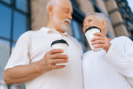 Low-angle view of beautiful gray-haired senior couple standing outside on summer day, holding coffee cups, enjoying each other company, loving looking at each other. Concept of happy retirementの写真素材
