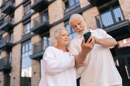 Low-angle view of happy senior couple standing embracing arm in arm on sunny day, using smartphone in front of modern brick apartment complex, exuding togetherness and love.の写真素材