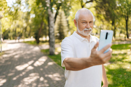 Portrait of handsome white-bearded senior man taking selfies on smartphone while walking on summer sunny park surrounded by lush greenery and trees. Happy elderly male capturing selfie in park.の写真素材