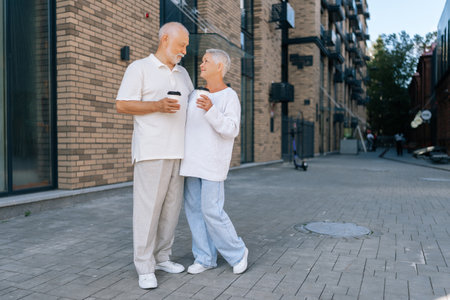 Full length portrait of loving senior couple enjoying coffee standing on city street, smiling talking, savoring togetherness in urban landscape on sunny day, on blurred background of building exteriorの写真素材
