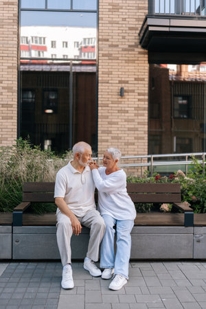 Full length vertical portrait of beautiful elderly couple enjoying happy moment sitting on city bench, embracing, warm talking and expressing love on summer day. Concept of retirement life.の写真素材