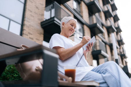 Shooting from below of pretty elegant senior woman peacefully taking notes sitting on city bench, exuding confidence and independence. Stylish happy elderly female taking notes on urban bench alone.の写真素材