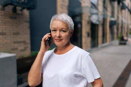 Over shoulder portrait of senior female talking on smartphone standing against urban backdrop with soft daylight, looking at camera. Happy older woman talking on smartphone with family.の写真素材