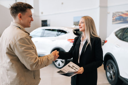 Portrait of female salesperson in business suit giving car keys to client, congratulating male buyer with purchase in auto showroom. Saleswoman and male customer shaking hands after successful dealの写真素材