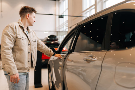 Male buyer in casual clothes choosing new car in dealership office, walking through showroom and looking at new car models with pensive expression. Concept of choosing and buying new auto at showroom.の写真素材
