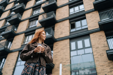 Shooting from below of fashionable redhead young woman walking through city drinking takeaway coffee and smiling while enjoying beautiful day. Stylish happy female standing with to-go coffee alone.の写真素材