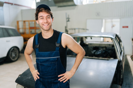 Cheerful male entrepreneur car repair shop standing on blurred background of cars under repair. Portrait of positive mechanic man in uniform and cap posing in auto repair workshop.の写真素材