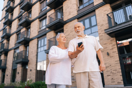 Shooting from below of cheerful elderly couple standing embracing on sunny day, using looking smartphone in front of modern brick apartment complex, exuding togetherness and love.の写真素材