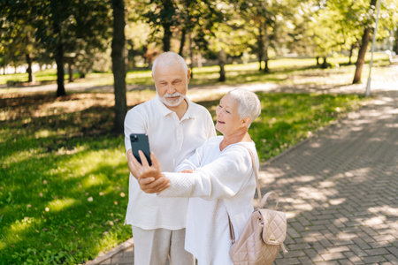 Cheerful gray-haired senior couple enjoying video call in summer park, surrounded by natural beauty...