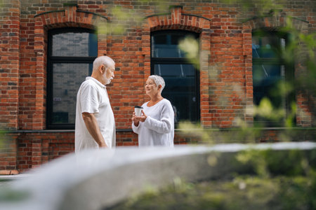 Remote wide shot of happy senior couple standing on city street and having coffee break, enjoying retirement and spending time together om warm sunny summer day. Concept of active agingの写真素材