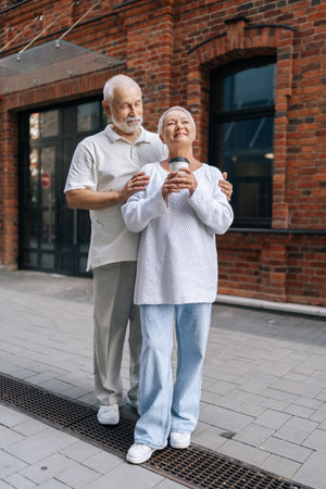 Full length vertical portrait of affectionate senior couple standing on city street, embracing and enjoying cup of coffee together, radiating happiness and contentment. Concept of active agingの写真素材