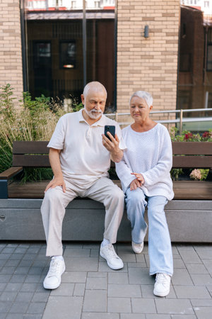 Full length vertical portrait of gray-haired elderly man and woman having online video call conversation, holding smartphone, looking to mobile phone camera, sitting on bench in courtyard of house.の写真素材