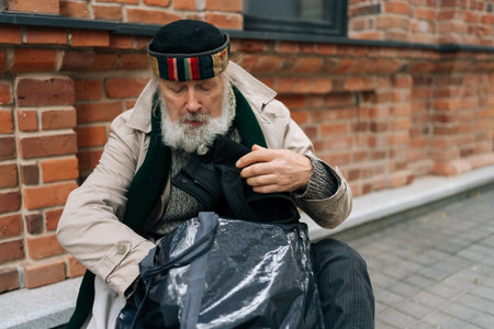 Portrait of homeless man with long white beard searching through belongings on cold day in city,...