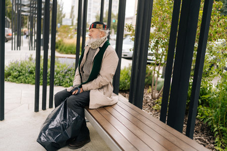 Pensive homeless elderly man in warm hat crouching outside alone on cold autumn day, face reflecting...
