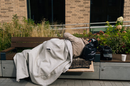 Back view of unrecognizable mature adult homeless person sleeping on urban bench, covered with...