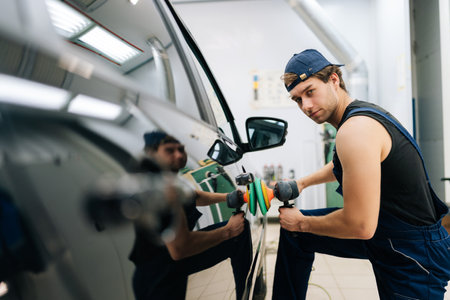 Portrait of confident mechanic male in uniform polish auto door using grinding unit in automobile...