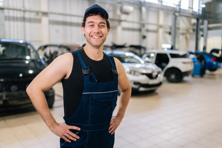 Medium shot portrait of cheerful technician male in uniform and cap standing posing auto service center, smiling looking at camera, enjoying time at workplace. Concept of professional car maintenance.の写真素材