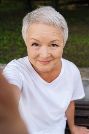 Cheerful elderly lady in park capturing selfie, appearing joyful, smiling looking at camera with...