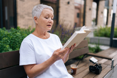 Side view of serene senior woman peacefully reading outdoors in city bench, embodying wisdom and experience. European gray-haired older female showcases happy retirement lifestyle.の写真素材