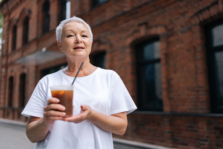 Portrait of happy senior woman with stylish short haircut, dressed in casual attire, savoring iced coffee while standing on city sidewalk, enjoying free time in retirement, looking at camera.の写真素材