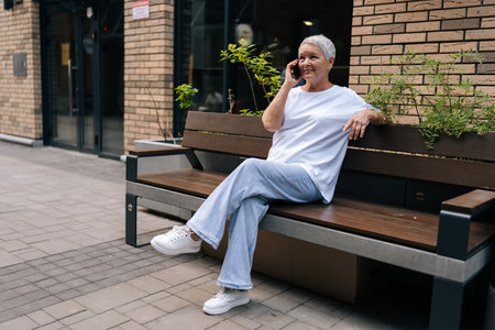 Wide shot of happy senior adult woman with gray hair sitting at bench on city street and talking on mobile phone. Attractive elderly female enjoys free time in retirement on European city.の写真素材