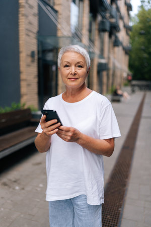 Vertical portrait of happy senior woman using typing smartphone while walking on street near modern...