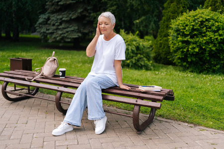 Unhappy elderly woman sitting in park bench feeling unwell, touching head, showing signs of pain and exhaustion, surrounded by nature. Reflects challenges of aging, mental health of senior healthcareの写真素材