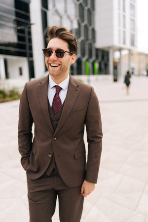 Vertical shot of joyful business man wearing stylish sunglasses and brown suit standing posing at city, smiling looking away, holding hands in pockets, exuding professionalism and leadership.の写真素材