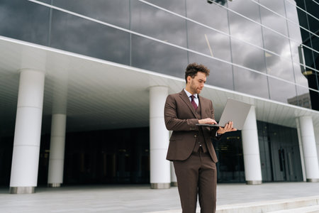 Handsome businessman in suit professional working on laptop standing outside modern office building,...